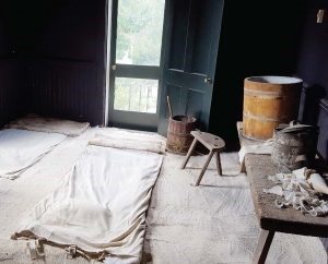 The interior of the Pest House in Lynchburg, Virginia. Not the barrel of linseed oil and limewater near the door to treat smallpox sores. The floor is covered with white sand, swept away daily, and sheets for patients to rest on. The black walls helped ease the eye stress and pain caused by the disease. (Photo by The Municipal.Com)